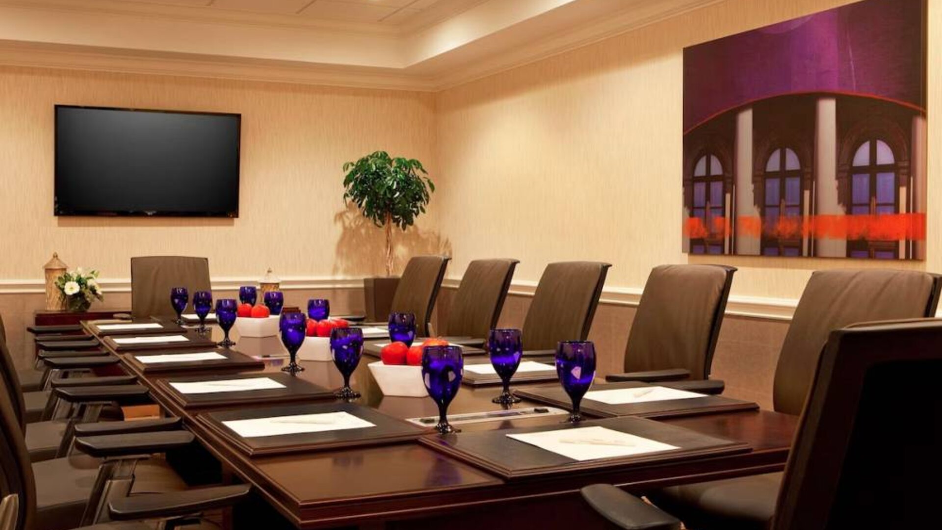 Meeting room in Sonesta Atlanta Airport North, furnished with long wooden table, a dozen chairs, and art and a widescreen television on the walls.