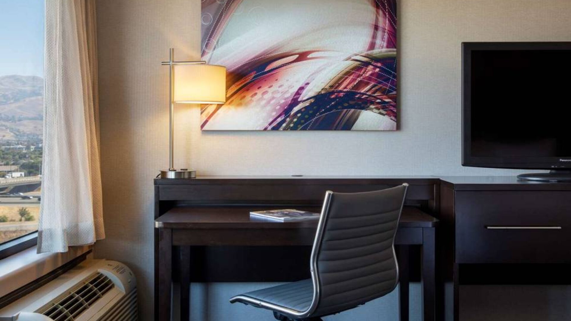 Sonesta San Jose guest room workspace, including desk, chair, lamp, and window, plus TV.