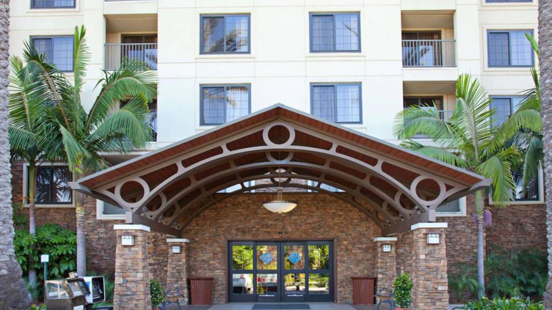 The hotel's exterior features a covered entrance, with potted plants and large trees and other greenery to either side.