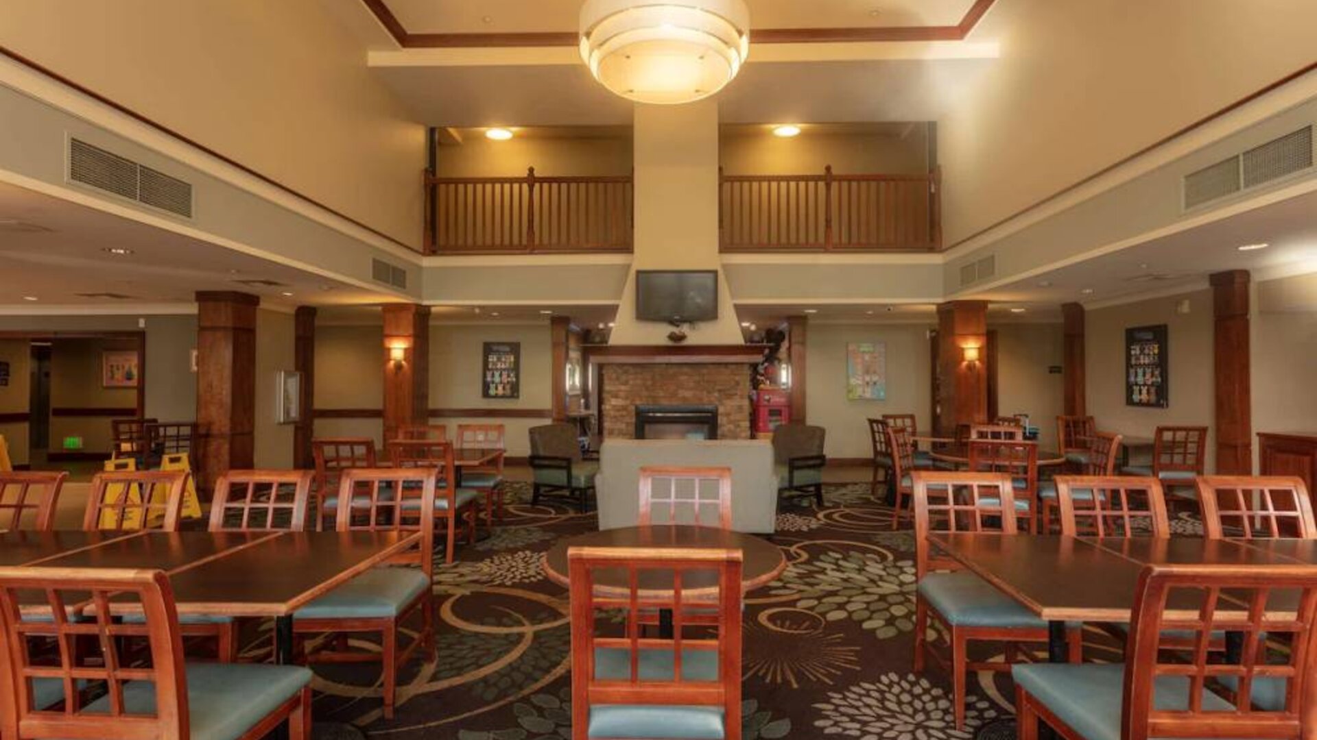 The hotel breakfast area has a high ceiling, wide range of table sizes, carpeted floor, and a wall-mounted TV.