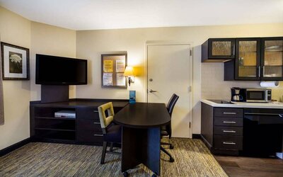 Sonesta Simply Suites St Louis Earth City guest room workspace, furnished with desk and chairs, with TV and kitchen nearby.