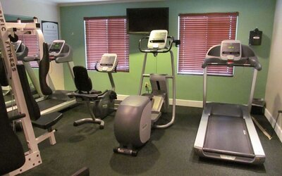 The hotel fitness center has two windows fitted with blinds, a wall-mounted TV, and  assorted exercise machines.