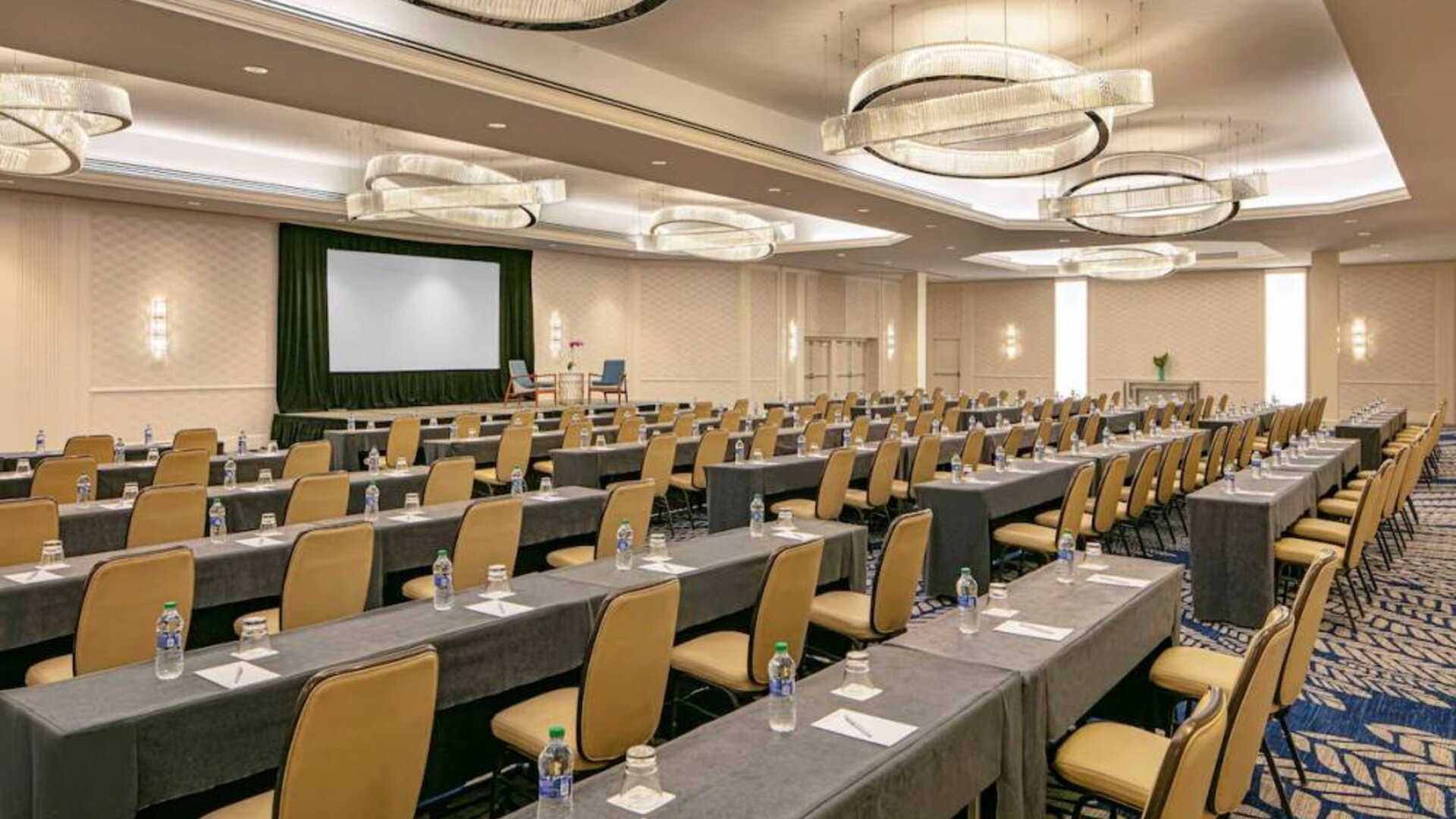 Hotel meeting room, furnished with tables and seating for dozens of attendees, arranged in a classroom style facing a large projector screen.