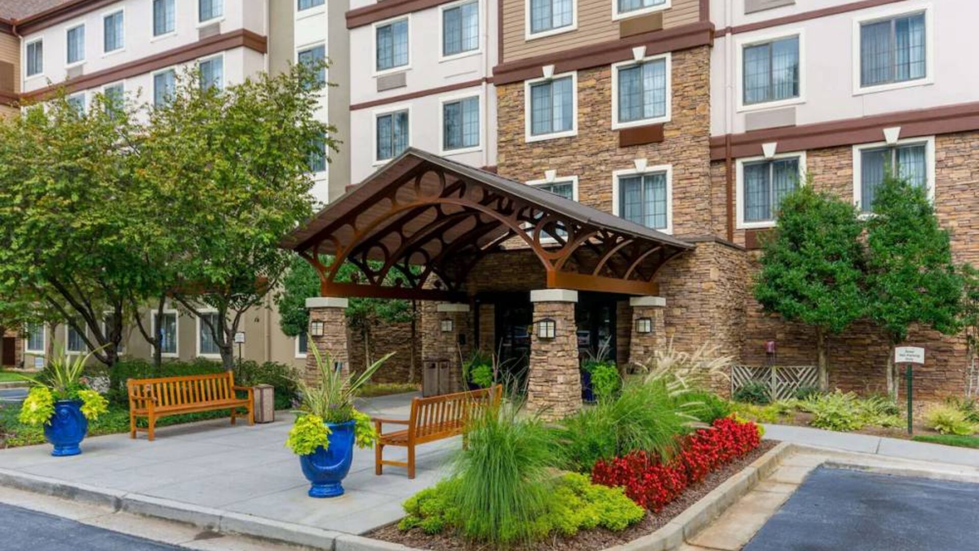 The hotel’s exterior has a pair of benches beside the covered entrance, with flowerbeds, bushes, and trees nearby.