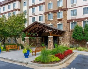 The hotel’s exterior has a pair of benches beside the covered entrance, with flowerbeds, bushes, and trees nearby.