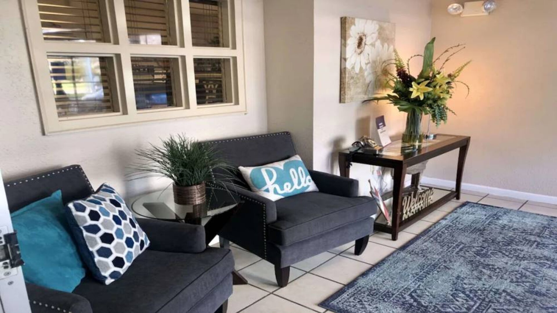 The hotel lobby is furnished with a pair of armchairs, a coffee table, art on the wall, and potted plants.