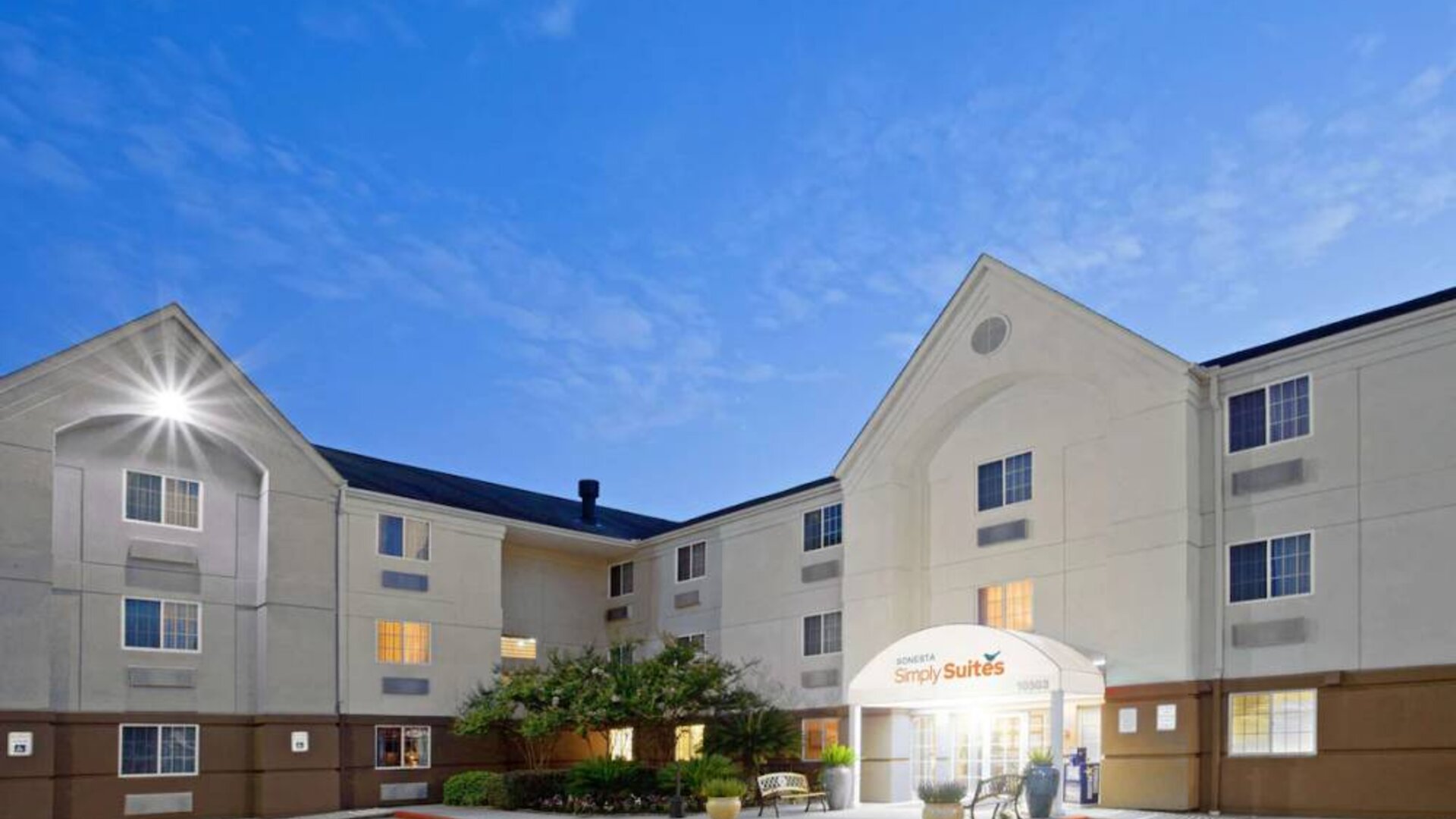The hotel’s exterior includes parking close to the front door, benches, potted plants, plus bushes, trees, and flowers.
