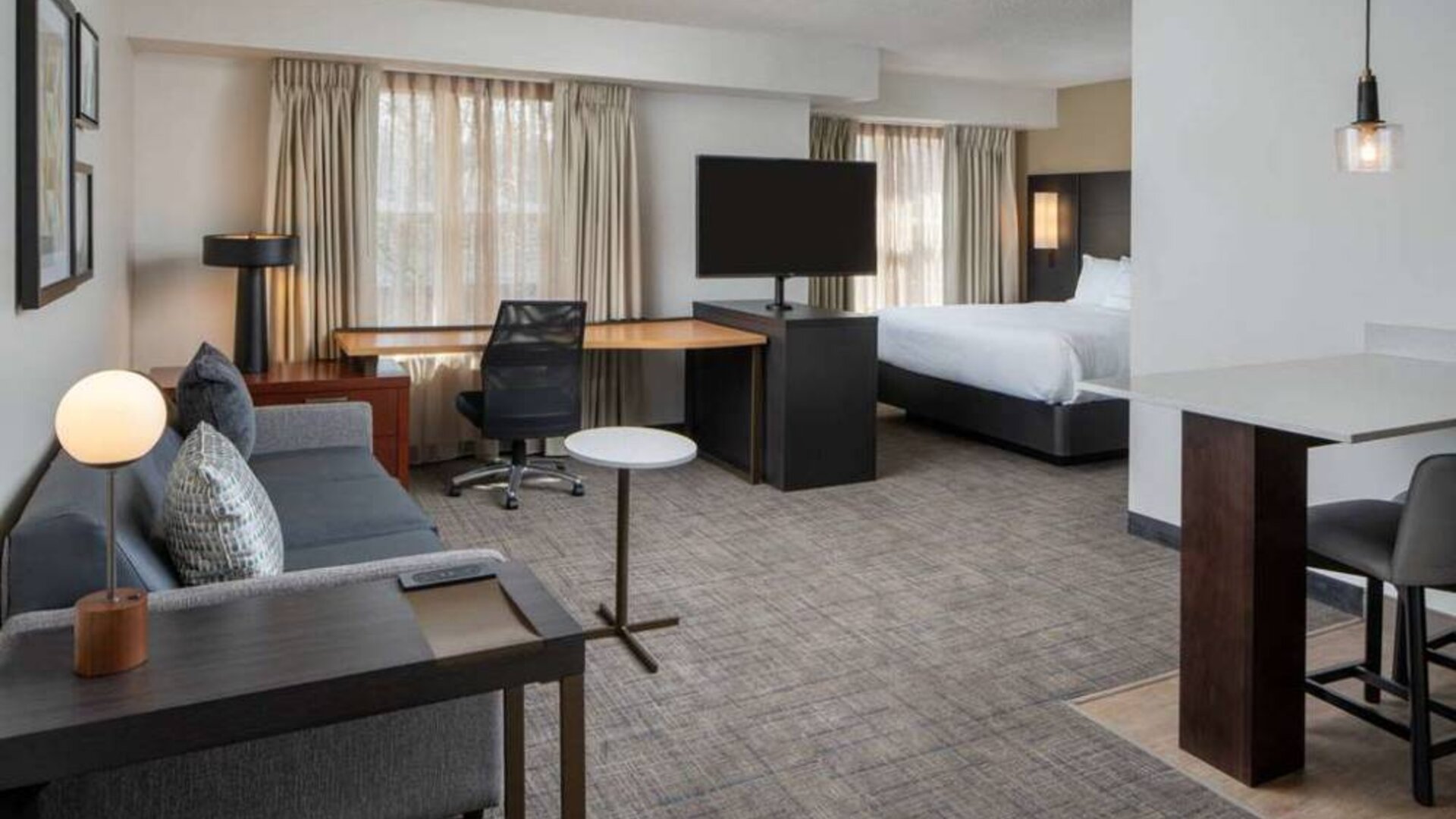 Sonesta ES Suites Fairfax Fair Lakes double bed guest room living area, furnished with sofa, chair, desk, and a large TV.