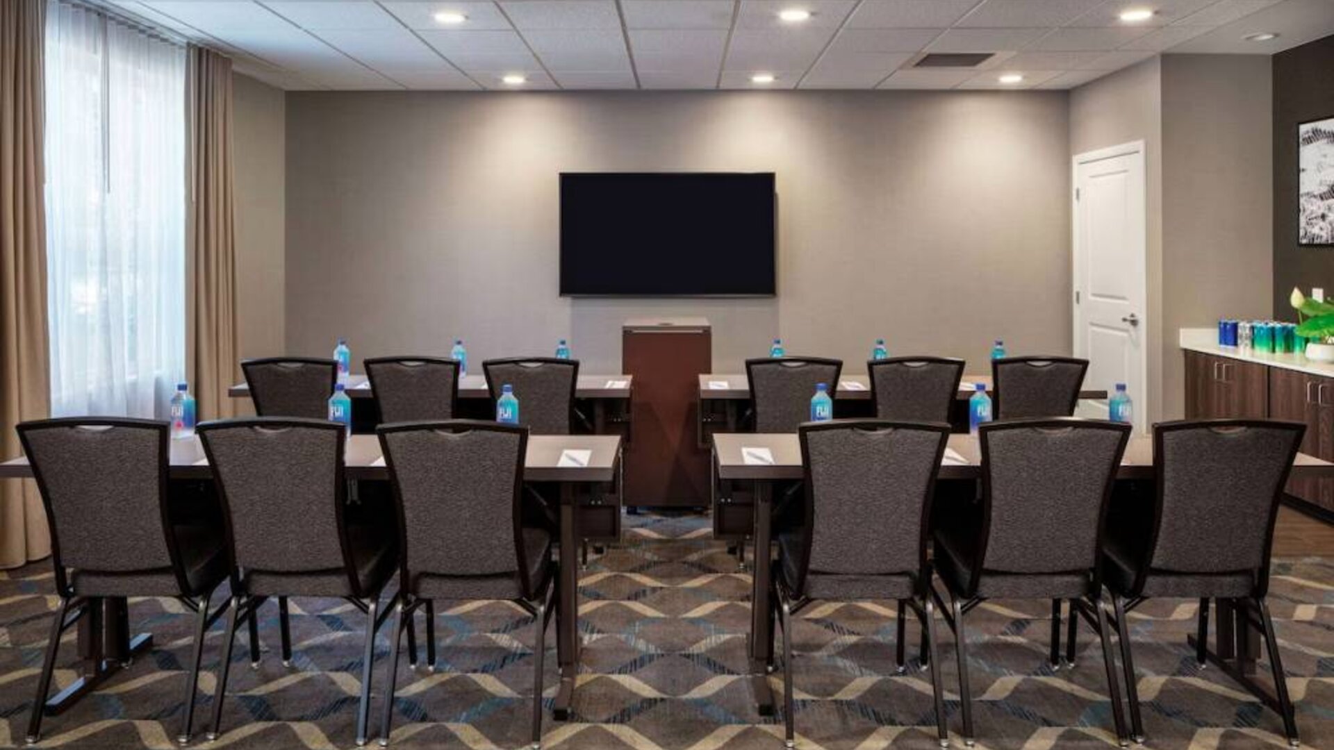 Hotel meeting room, with tables and twelve seats facing a lectern and wall-mounted TV.