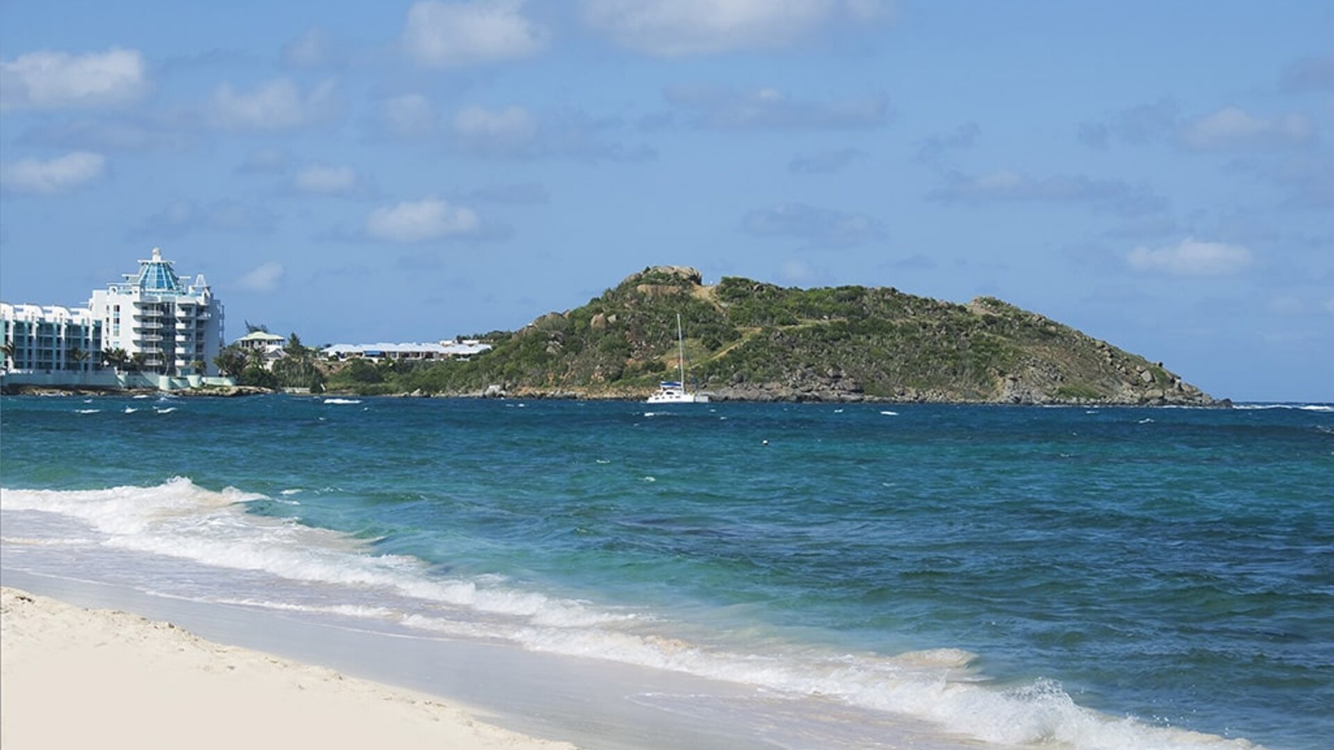 Waves coming on shore with view of resort buildings and mountainous island in distance. 