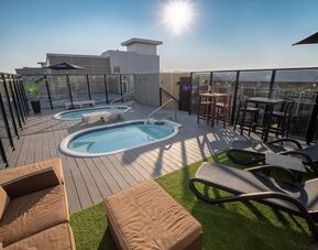 Rooftop deck with two hot tubs, lounge seating, high-top tables, and glass safety railing in daylight. Hotel Clique Calgary Airport.