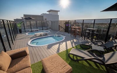 Rooftop deck with two hot tubs, lounge seating, high-top tables, and glass safety railing in daylight. Hotel Clique Calgary Airport.