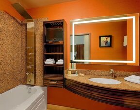 Bathroom with soaking tub, rainfall shower panel, illuminated mirror, granite vanity, and open shelving. Hotel Clique Calgary Airport.