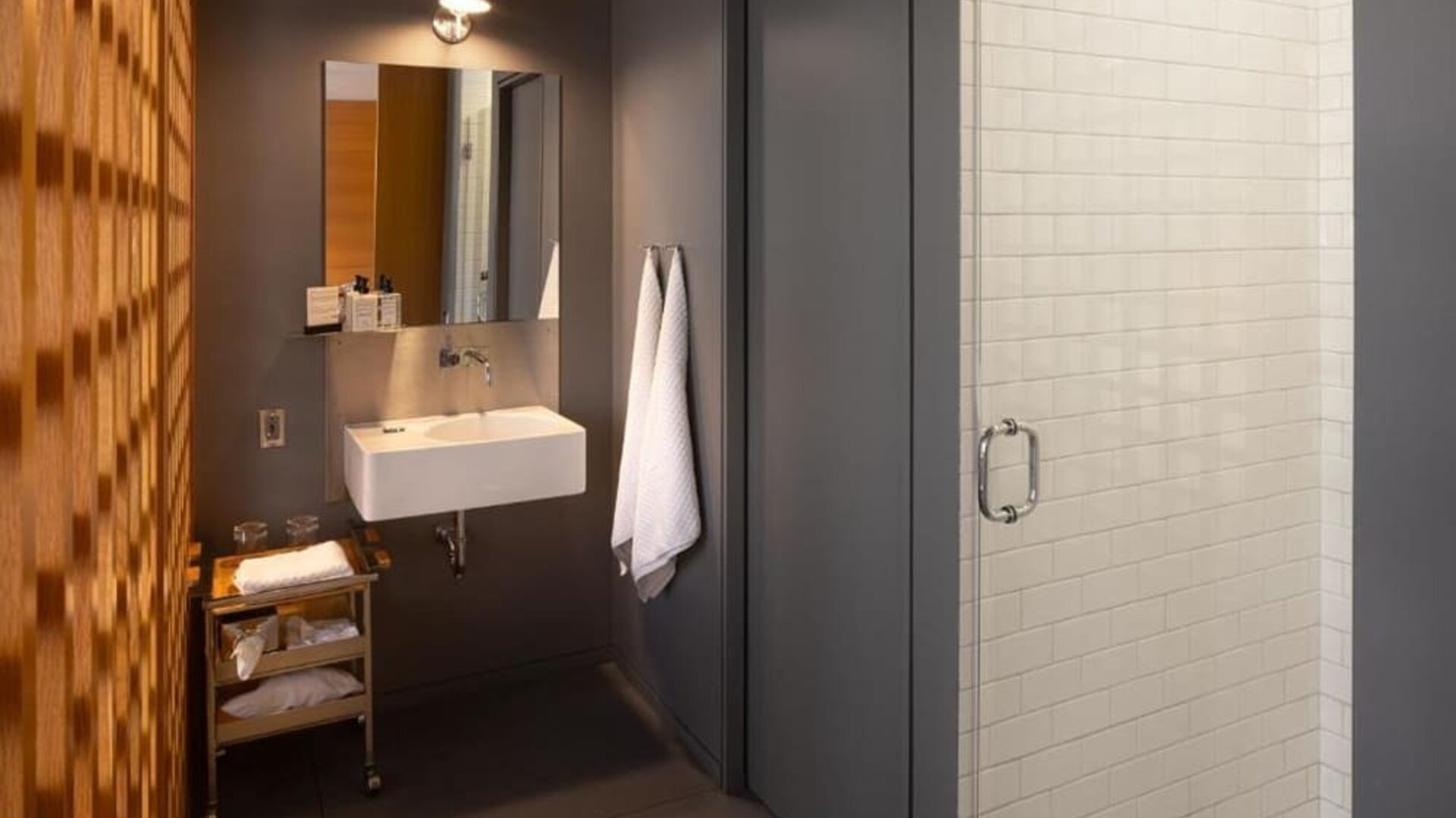 Modern bathroom with glass walk-in shower, floating vanity, open shelving, and white subway tile walls. Hotel Gault.