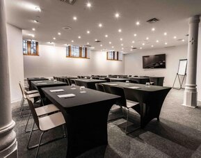 Meeting room with classroom-style table layout, presentation screen, flip chart, and overhead lighting. Hotel Gault.