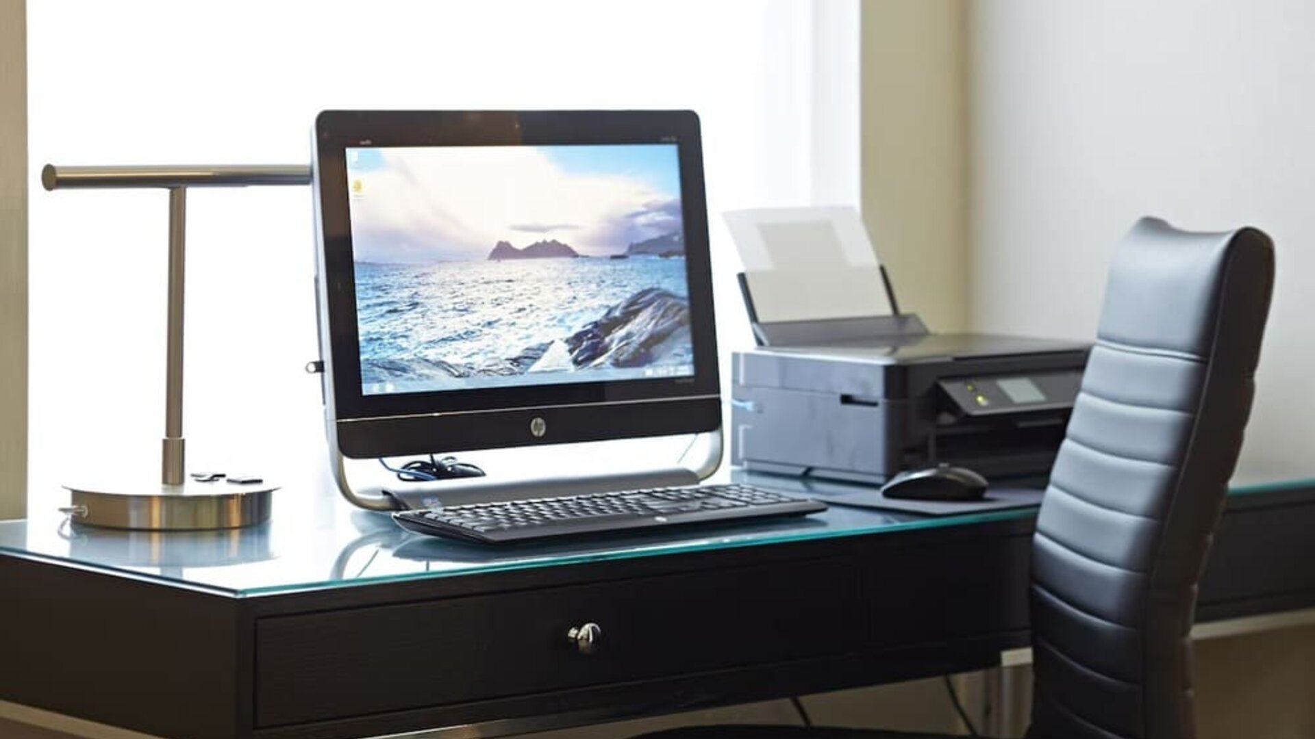 Business center workstation with desktop computer, printer, glass desk, and black leather office chair at Comfort Inn Boucherville.