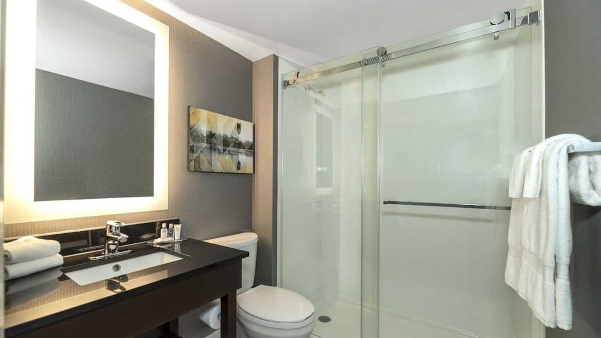 Modern bathroom with illuminated mirror, walk-in glass shower, black vanity, and fresh towels at Comfort Inn Boucherville.