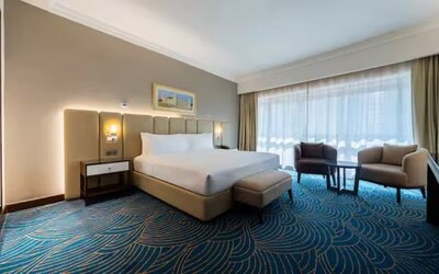 Spacious king room with floor-to-ceiling windows, lounge seating, patterned carpet, and natural daylight.