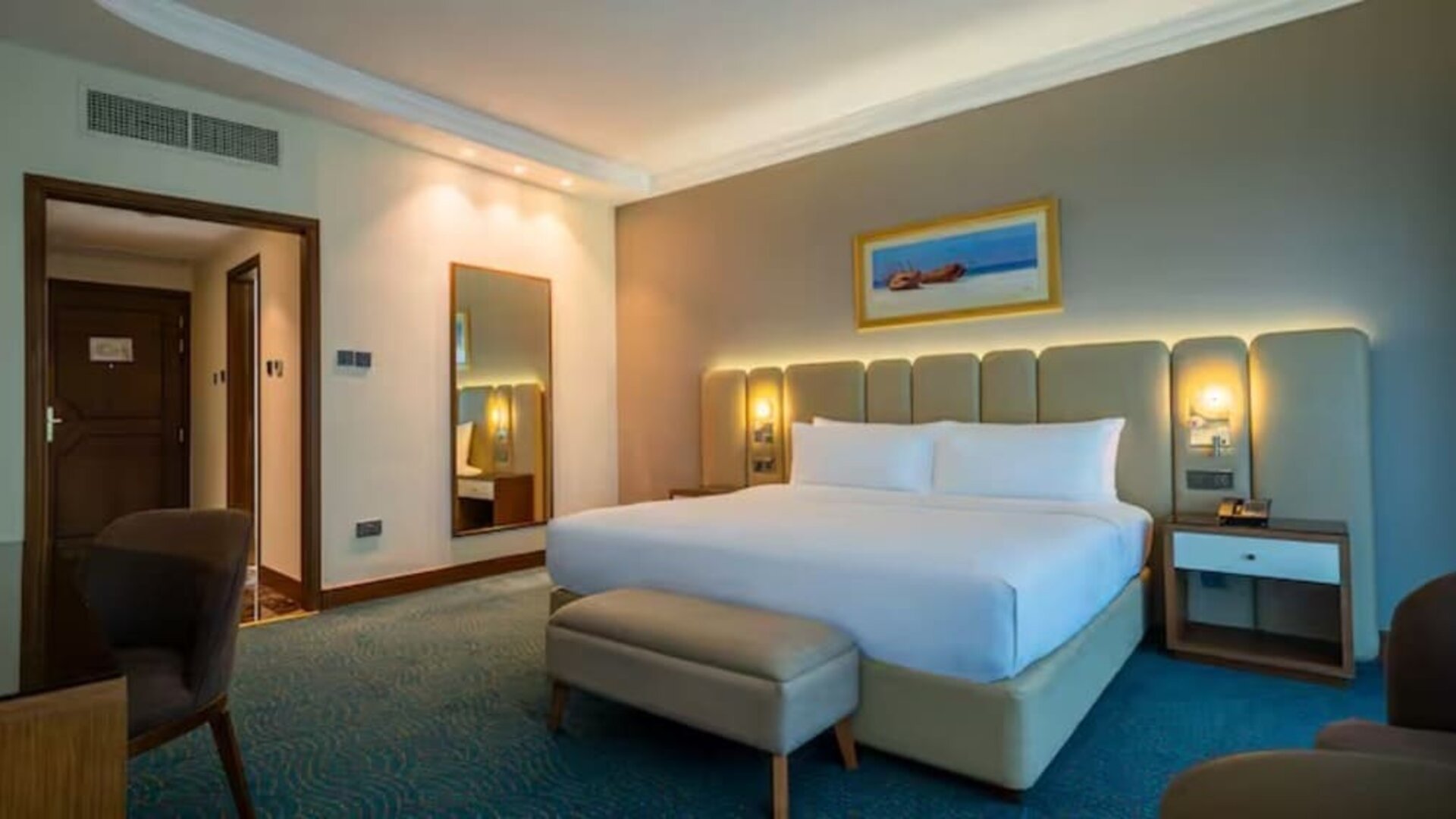 Bright guest room featuring a king bed, upholstered headboard, bedside lighting, seating area, and workspace.