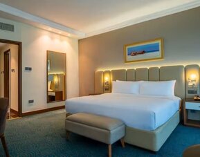 Bright guest room featuring a king bed, upholstered headboard, bedside lighting, seating area, and workspace.