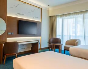 Contemporary guest room with two twin beds, large window, seating area, wall-mounted television, and workspace.