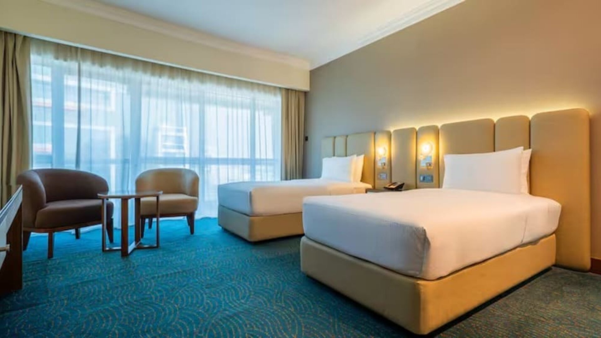 Comfortable twin-bed room with upholstered headboards, sitting area, floor-to-ceiling curtains, and soft ambient lighting.