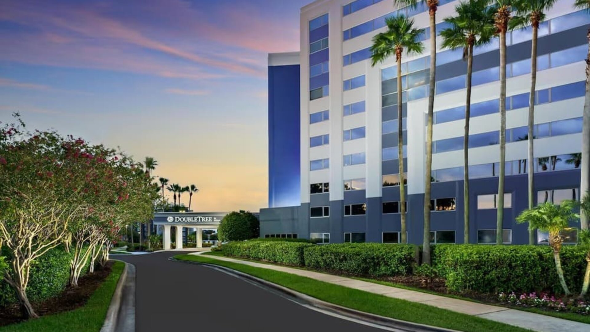 Palm-lined hotel entrance with landscaped gardens, curved driveway, and multi-story exterior at sunset.