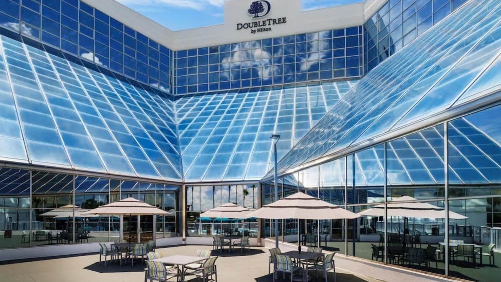 Open-air terrace with patio tables, umbrellas, and glass hotel façade reflecting blue skies.