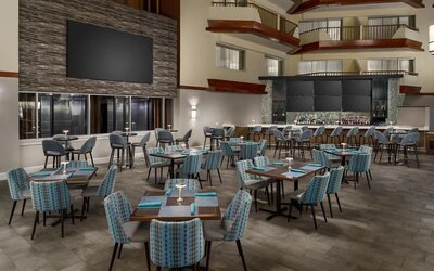 Spacious hotel restaurant with dining tables, upholstered chairs, atrium-style architecture, and adjacent bar area.
