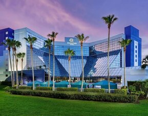 Glass-fronted hotel exterior surrounded by palm trees and manicured lawns under a colorful evening sky.