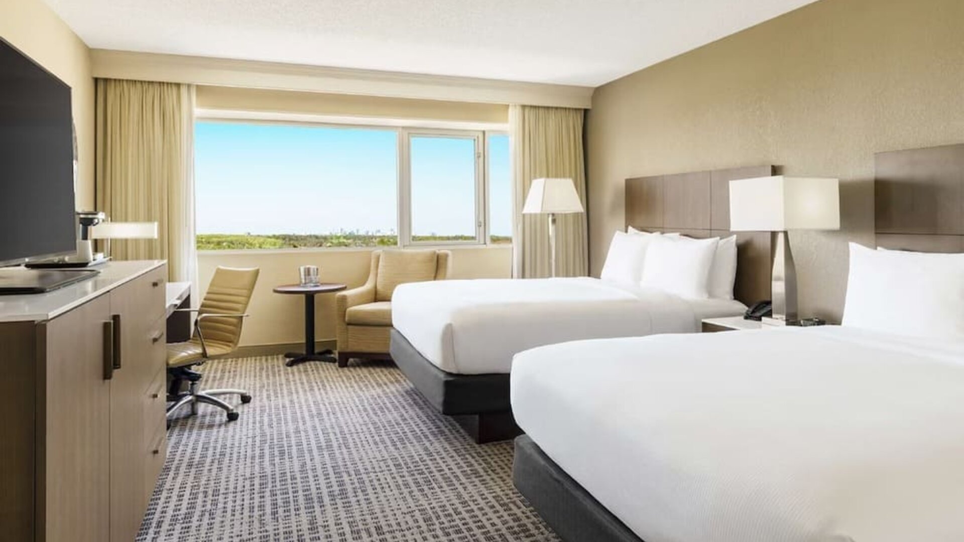 Guest room with two double beds, work desk, lounge chair, large window, and bright contemporary furnishings.