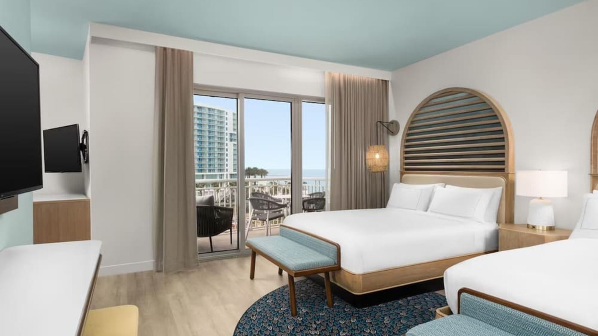 Guest room with two beds, patterned rug, balcony access, and views toward the beach and nearby buildings.