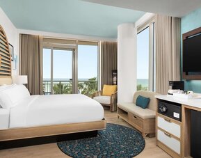 Contemporary guest room with king bed, seating area, large TV, and balcony offering partial ocean views.