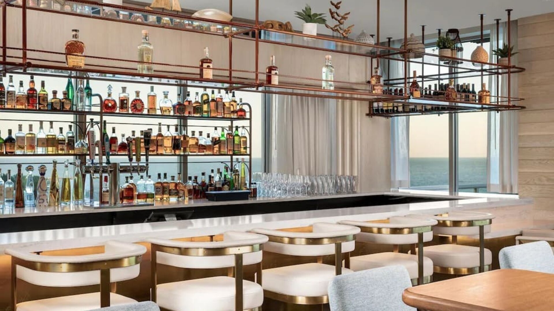 Elegant ocean-view bar with marble countertop, cushioned seating, hanging shelves, and extensive liquor display.