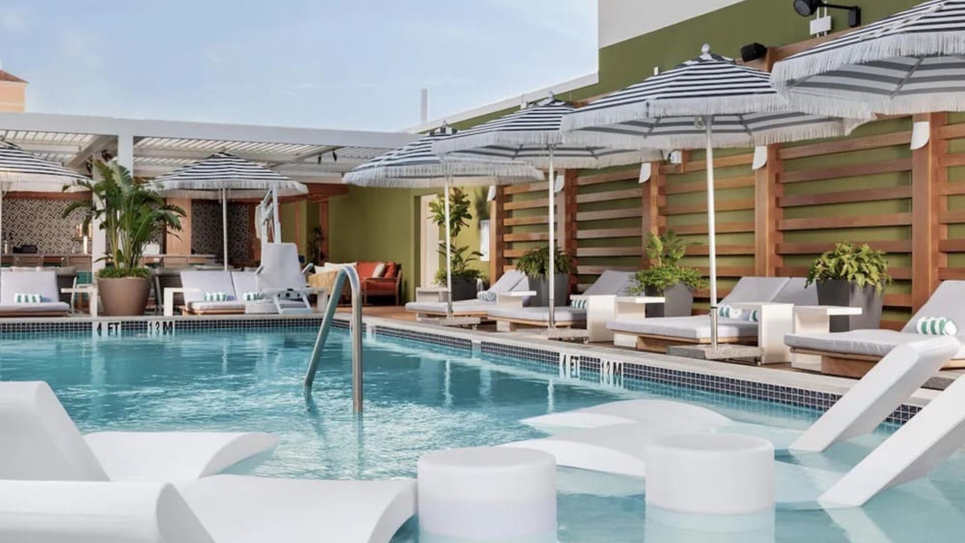 Rooftop pool deck with in-water loungers, striped umbrellas, ocean views, and comfortable relaxation areas.