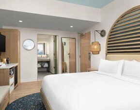 Bright guest room with king bed, wood accents, wall-mounted TV, and open vanity area leading to bathroom.