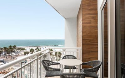 Balcony with round table and four woven chairs overlooking white-sand beach, palm trees, and turquoise ocean.