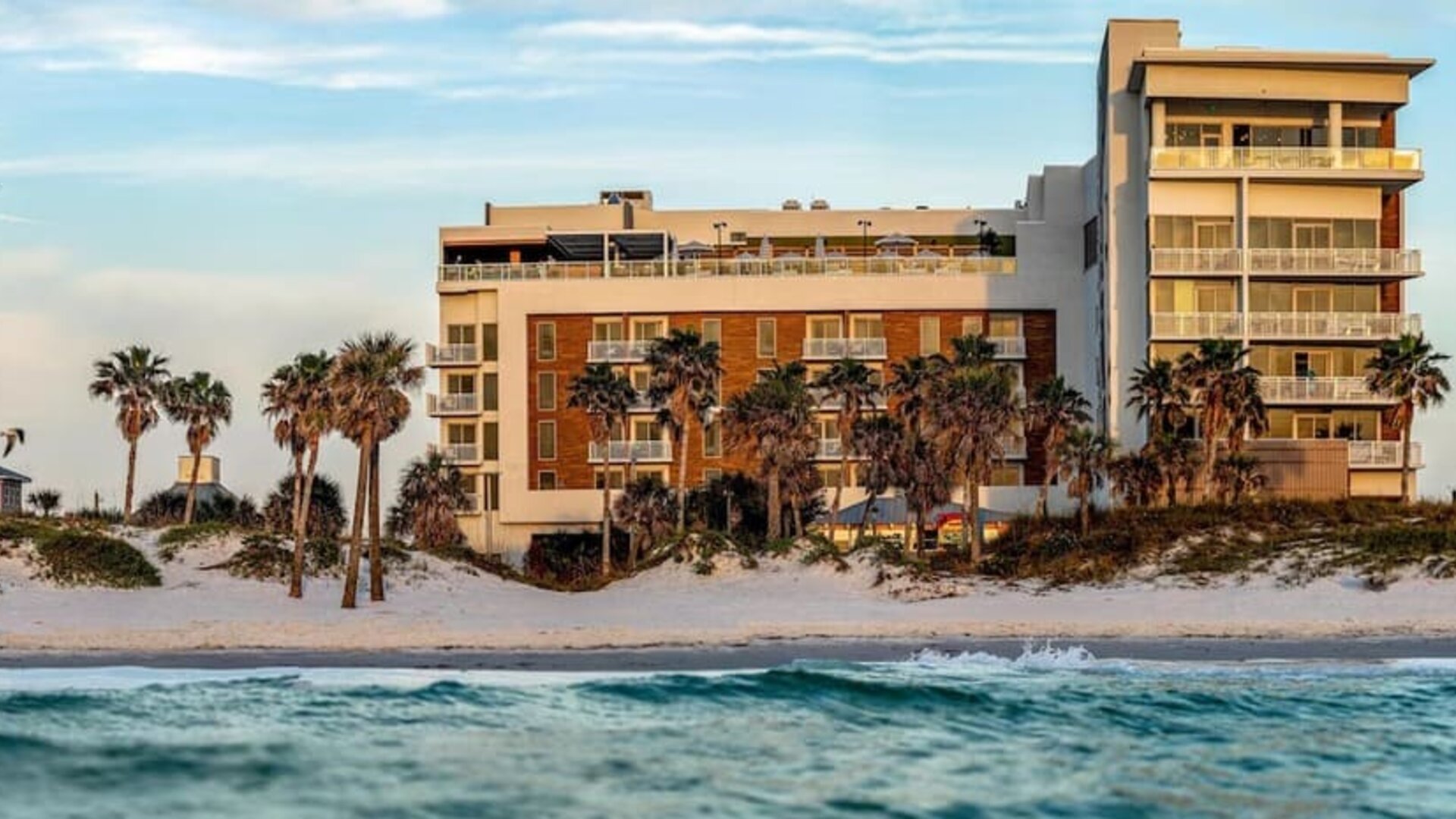 Beachfront hotel exterior framed by palm trees, white sand dunes, and gentle waves along the shoreline.