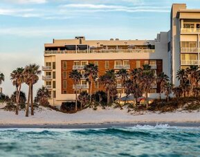 Beachfront hotel exterior framed by palm trees, white sand dunes, and gentle waves along the shoreline.