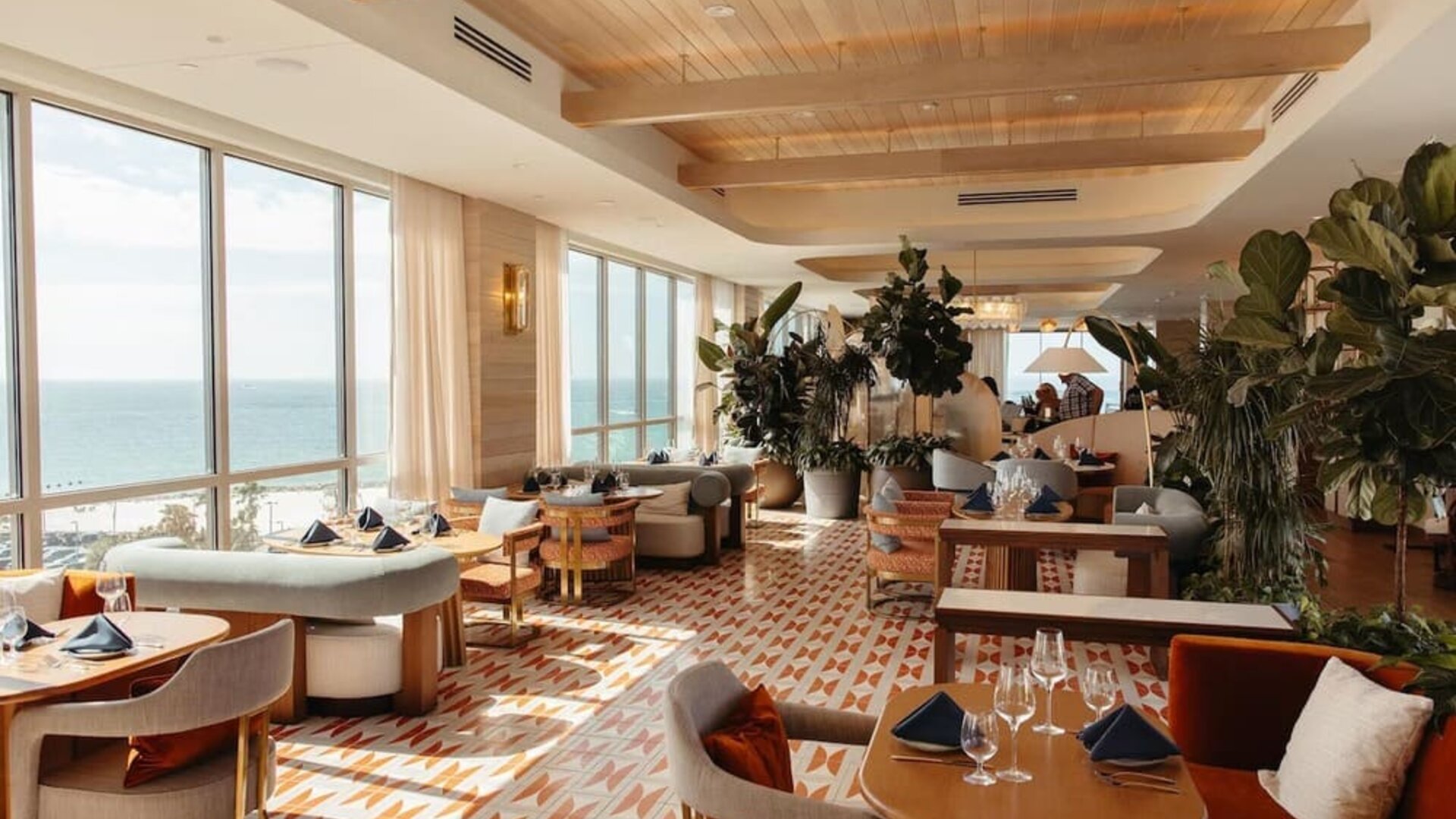 Upscale restaurant and lounge with ocean-facing windows, stylish seating, lush plants, and natural daylight.
