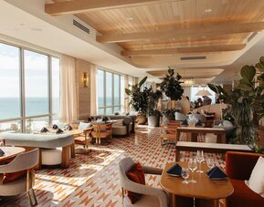 Upscale restaurant and lounge with ocean-facing windows, stylish seating, lush plants, and natural daylight.