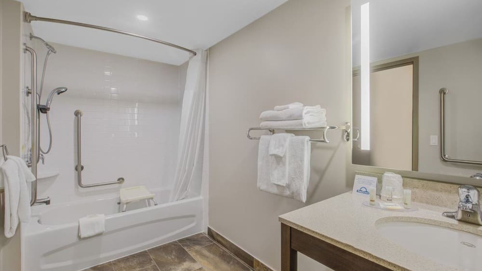Accessible guest bathroom with bathtub, grab bars, shower seat and illuminated vanity at Days Inn & Suites By Wyndham Lindsay.