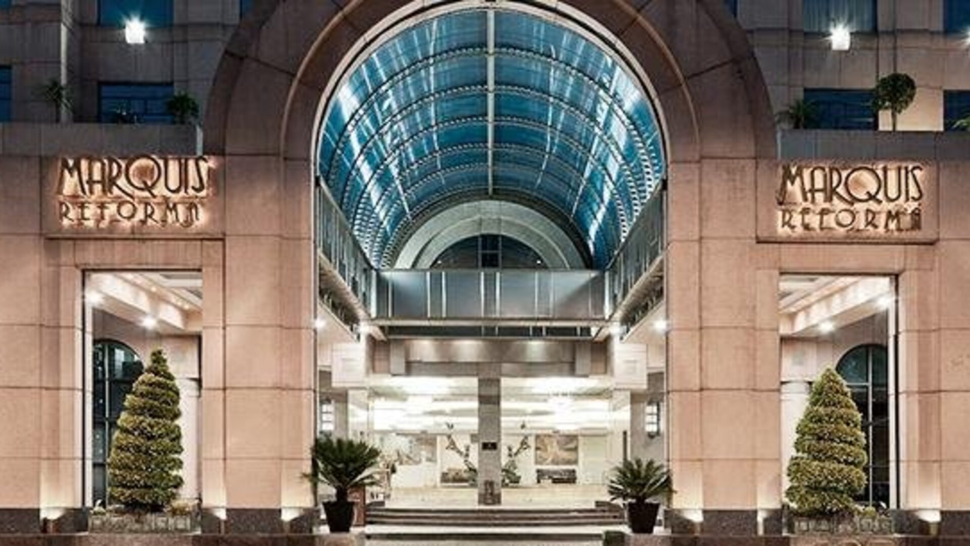 Main entrance of Hotel Marquis Reforma featuring an illuminated arched glass canopy and landscaped planters.