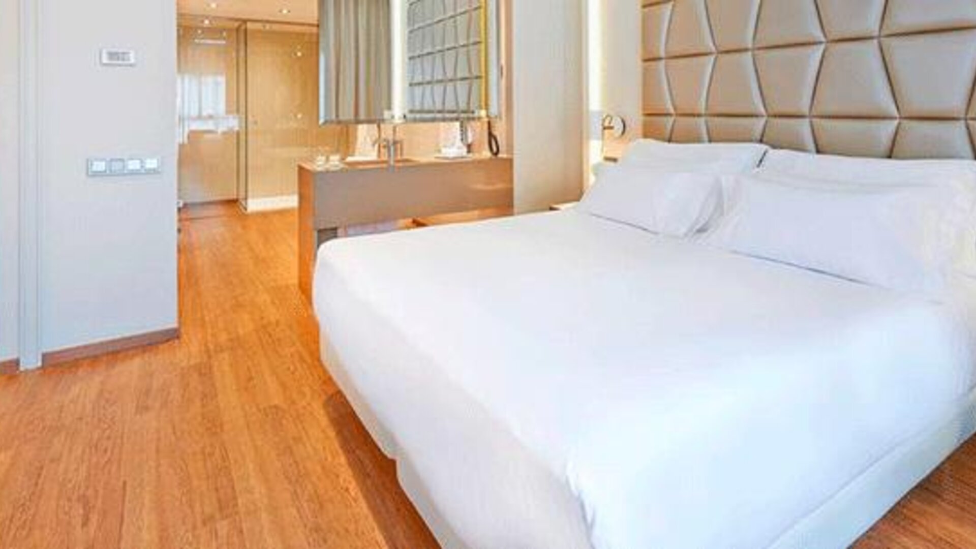 Contemporary guest room featuring a large bed, geometric upholstered headboard, wood flooring and open bathroom area at NH Collection Barcelona Pódium.
