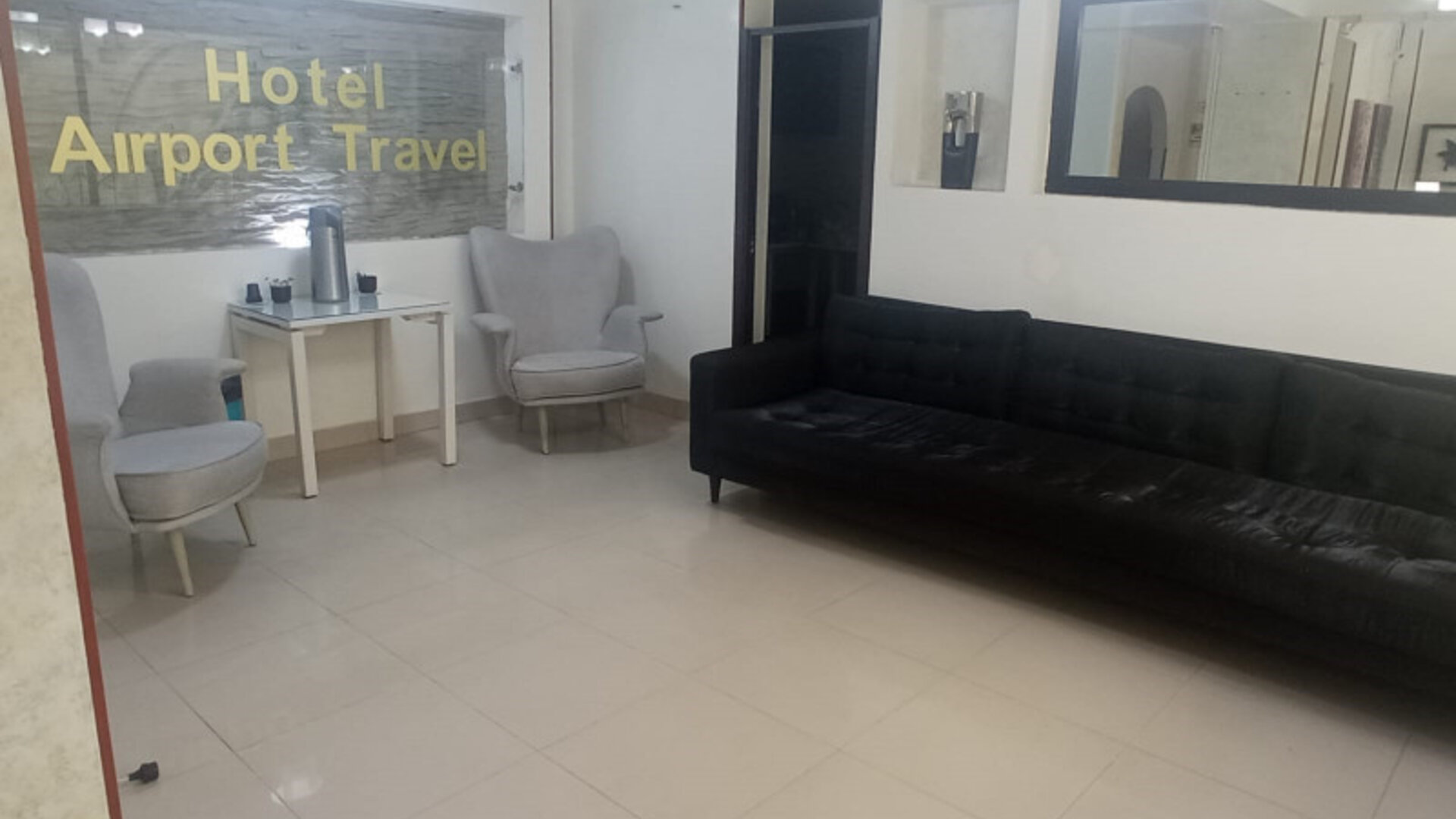 Lobby seating area featuring a black sofa, armchairs, large wall mirror and refreshment table at Hotel Airport Travel.