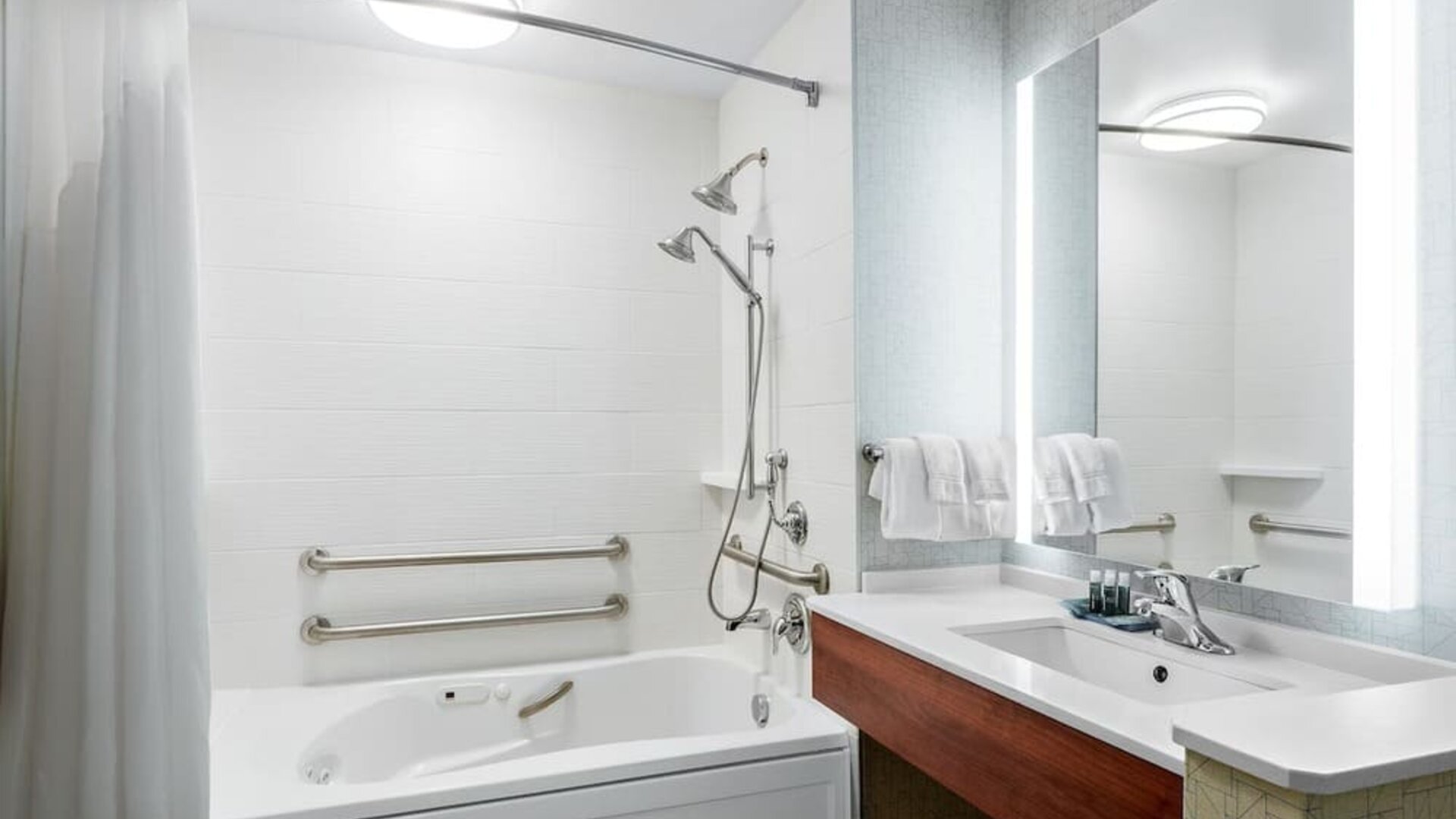 Accessible bathroom featuring a bathtub with grab bars, an adjustable showerhead and a modern vanity with an illuminated mirror at Holiday Inn Express & Suites Ontario.