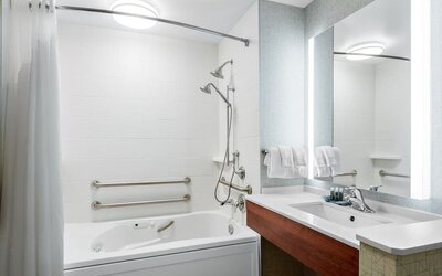 Accessible bathroom featuring a bathtub with grab bars, an adjustable showerhead and a modern vanity with an illuminated mirror at Holiday Inn Express & Suites Ontario.
