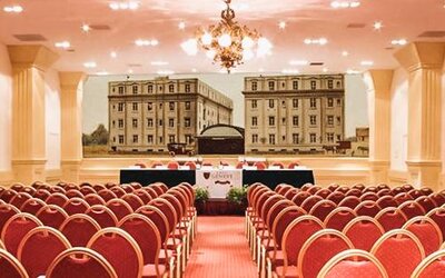 Large conference hall with rows of red chairs, a presentation stage, chandeliers, and a decorative mural backdrop at Hotel Geneve.