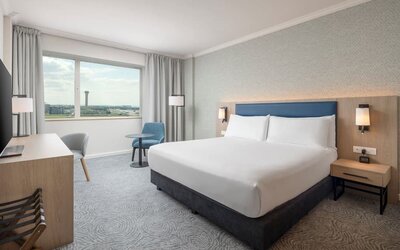 Modern king guest room with bed, large television, long work desk, armchairs, side table and floor-to-ceiling curtains.