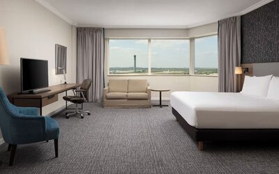 Spacious hotel room with king bed, sofa, work desk, television, armchair and panoramic windows overlooking the airport.